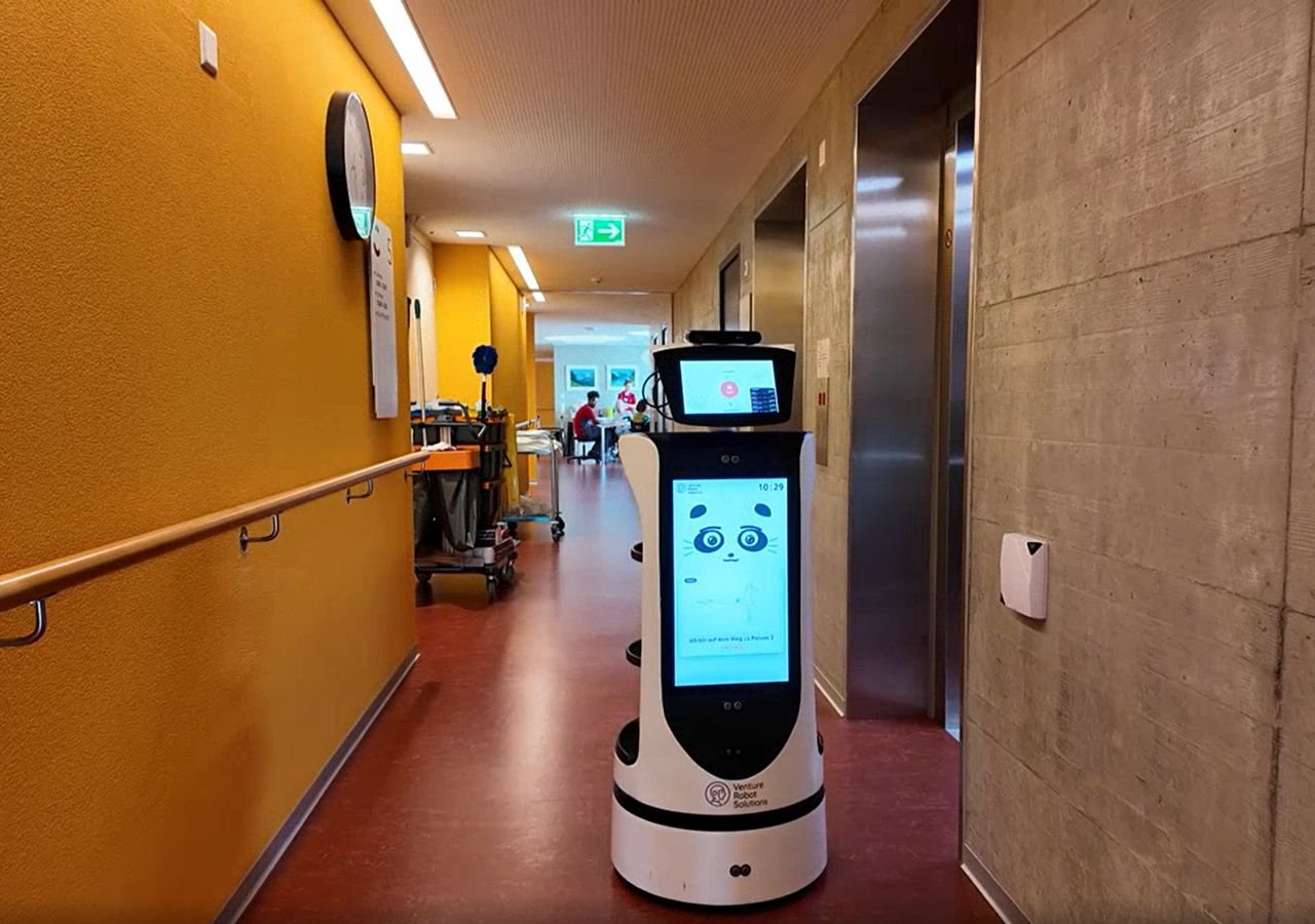 The robot running a delivery mission in a Haus Tabea corridor, its screen announcing the resident it is on the way to